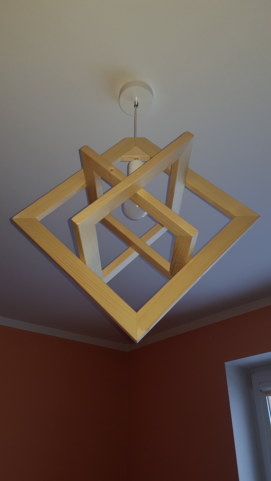 Nowoczesna lampa wisząca z jasnego drewna, składająca się z kwadratowych ram, zawieszona na białym suficie. Widoczna żarówka i fragment pomarańczowej ściany.