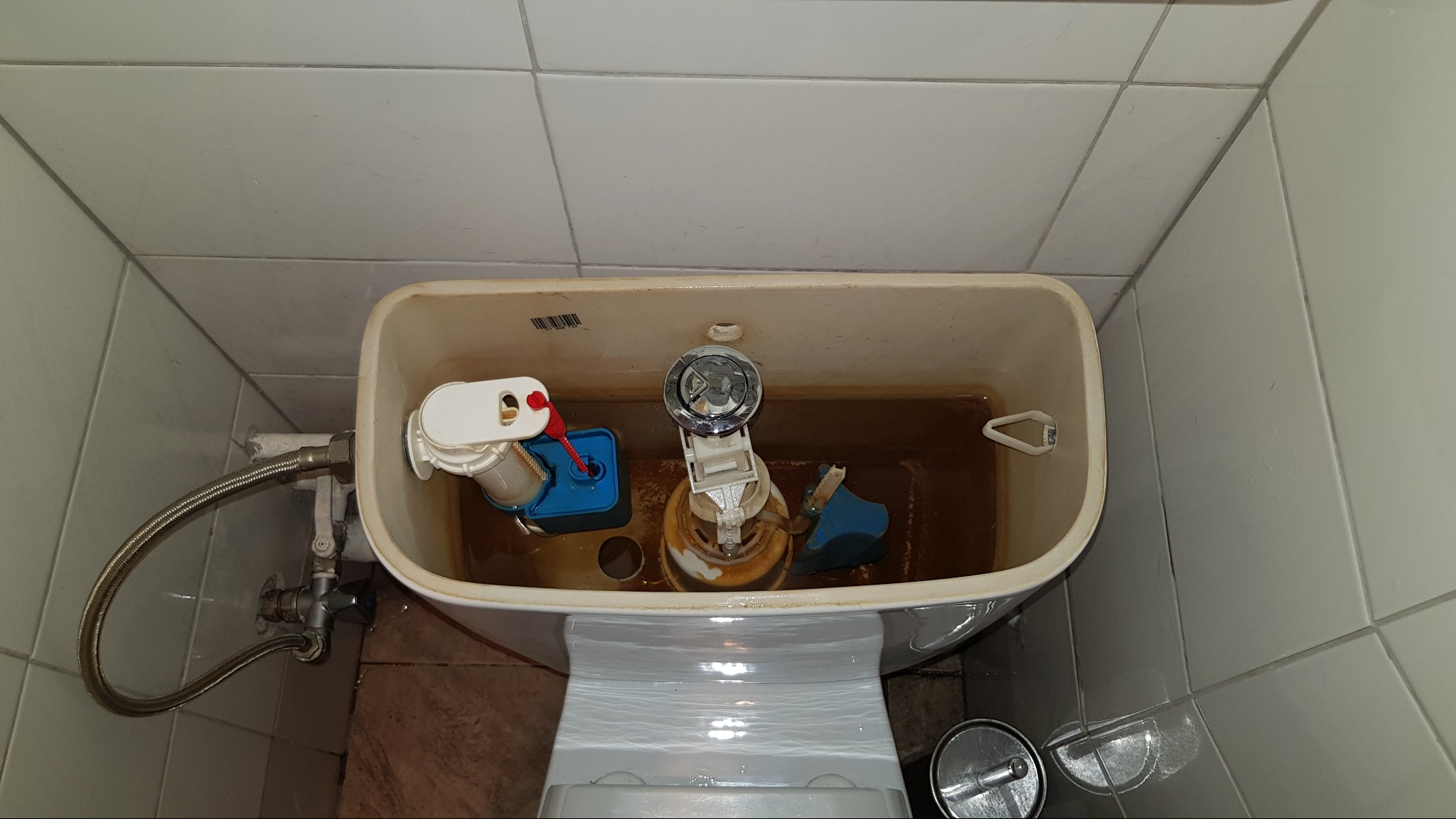 Wnętrze spłuczki toalety z widocznymi elementami mechanicznymi, woda o brązowym zabarwieniu, białe płytki w tle. Ujęcie z góry.