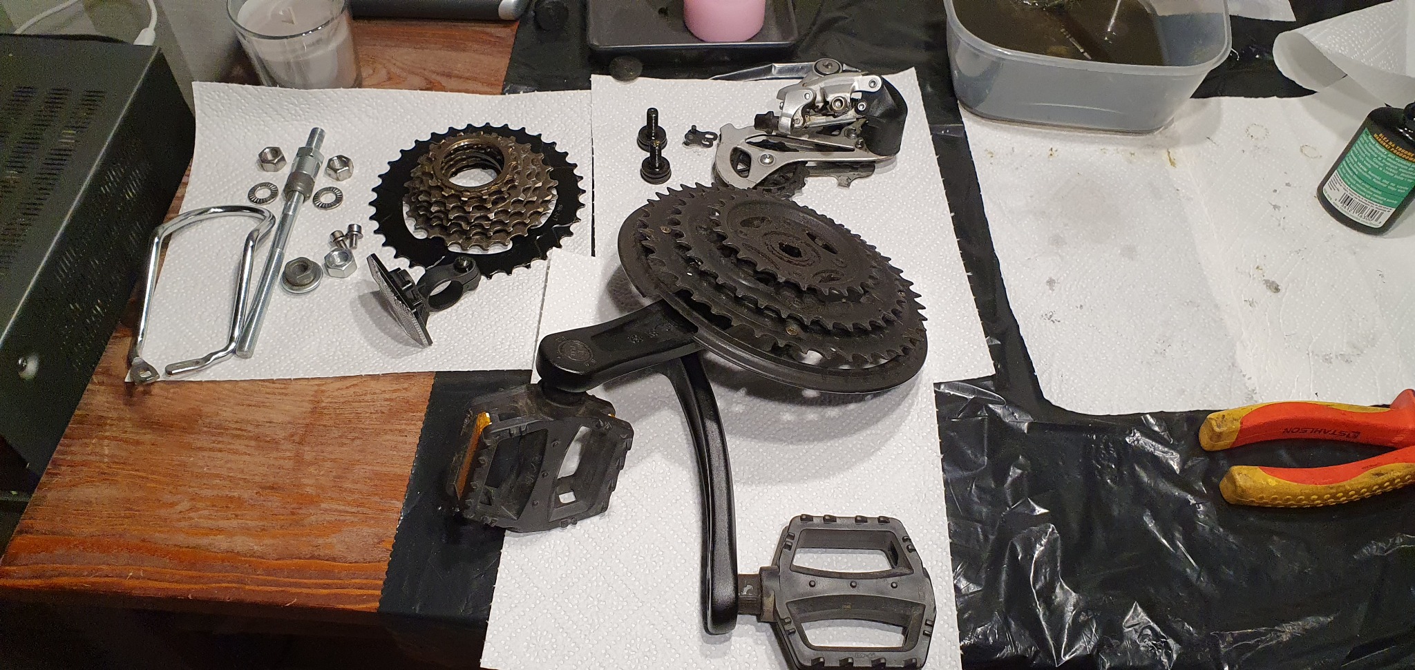 Rozłożone części roweru na stole: przerzutka, kaseta, korba z pedałami, śruby, uchwyt na bidon i narzędzia. Przygotowanie do serwisu lub naprawy roweru.