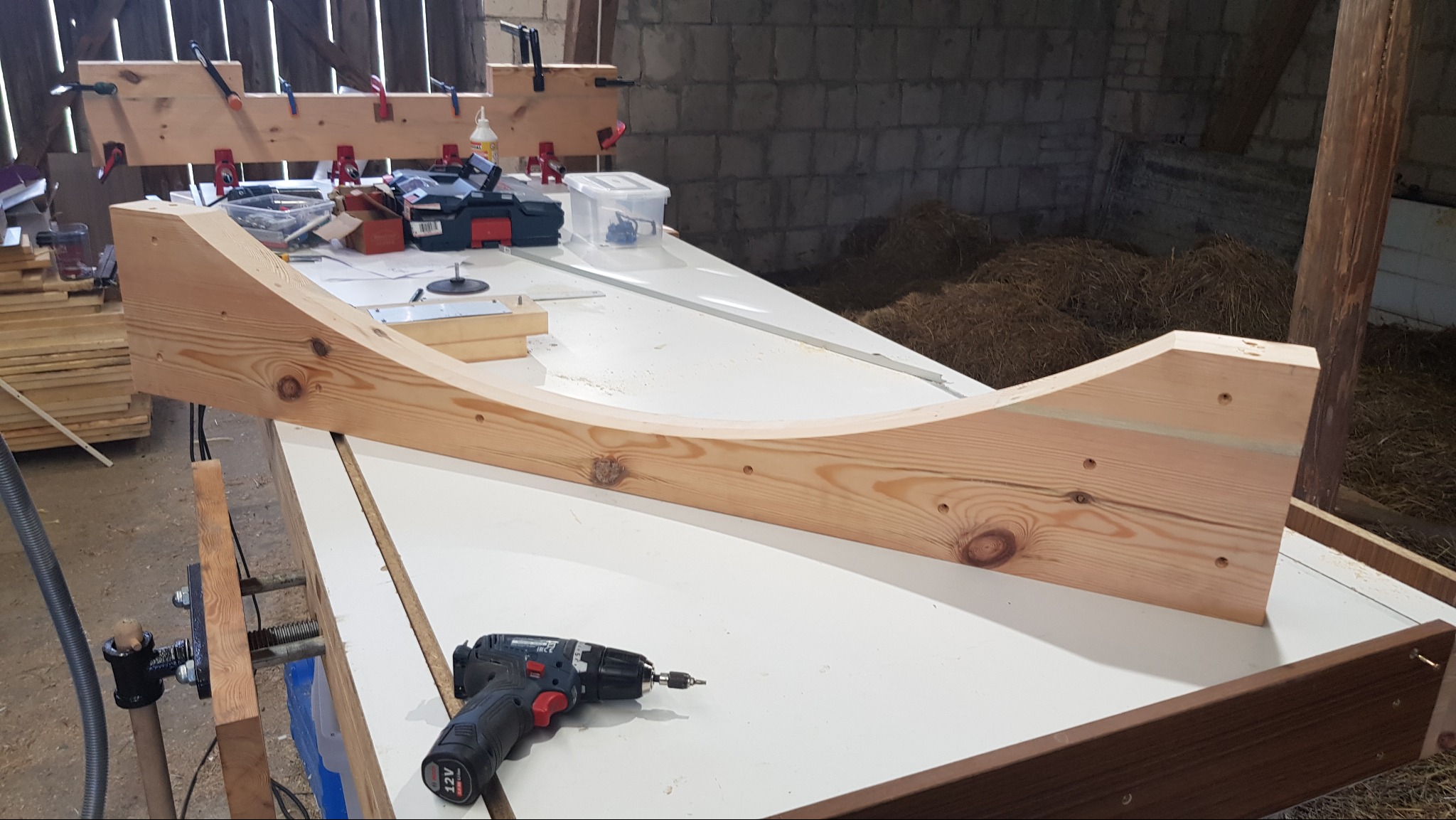 Element z jasnego drewna o zakrzywionym kształcie na stole warsztatowym, obok wiertarka. W tle narzędzia i bele siana. Detal stolarski w surowym warsztacie.