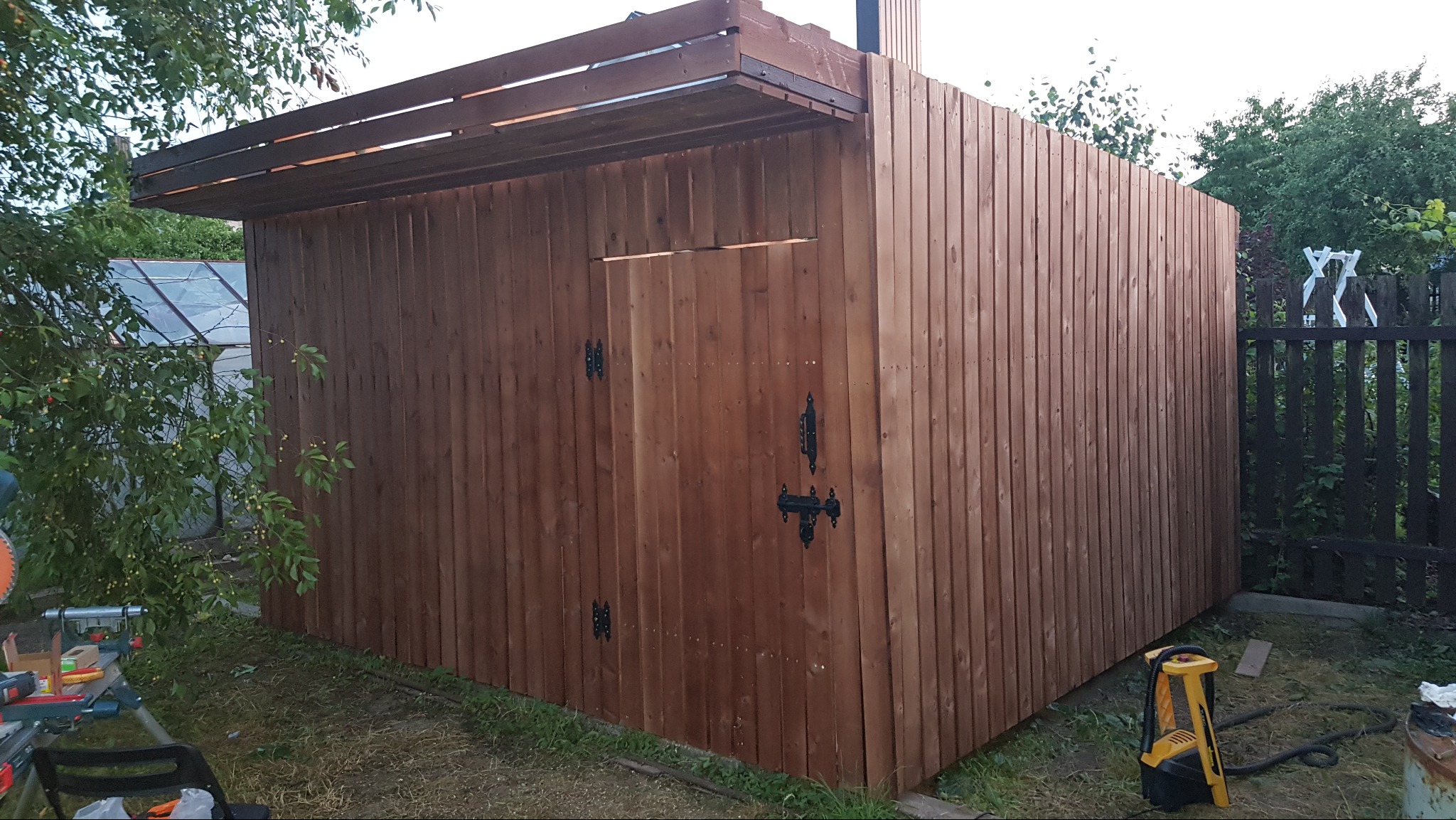 Drewniana konstrukcja z zadaszeniem, prawdopodobnie szopa lub mały budynek gospodarczy, wykonana z pionowo ułożonych desek, z czarnym zamkiem na drzwiach.