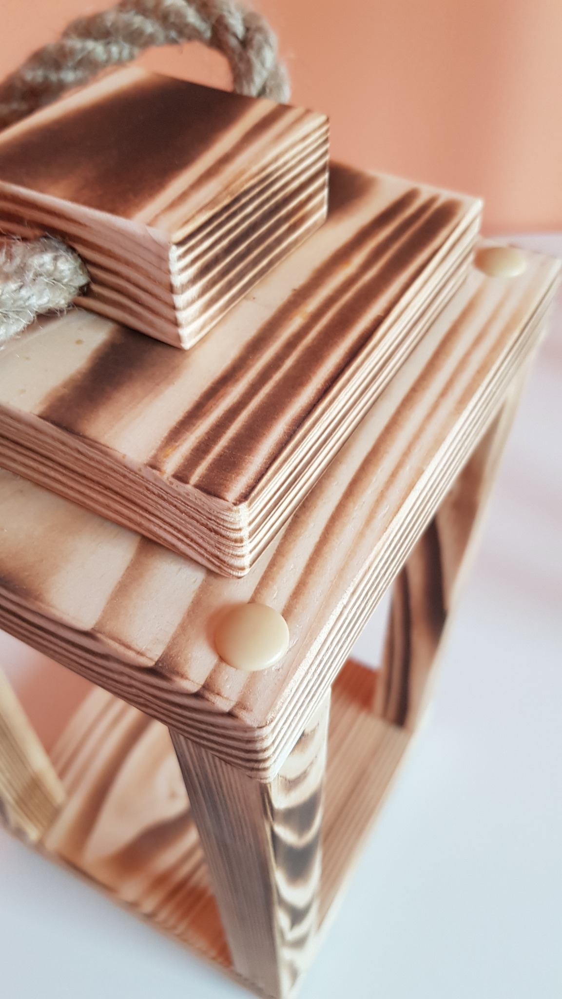 Drewniana konstrukcja z wypalanymi wzorami, połączona sznurkiem. Widoczne warstwy drewna i kremowe elementy mocujące. Ujęcie z góry, na jasnym tle.