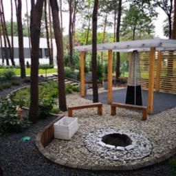 Ogród z paleniskiem otoczonym kamieniami, drewnianymi ławkami i pergolą. Widoczne drzewa i nowoczesny budynek w tle. Czarna kora i roślinność.