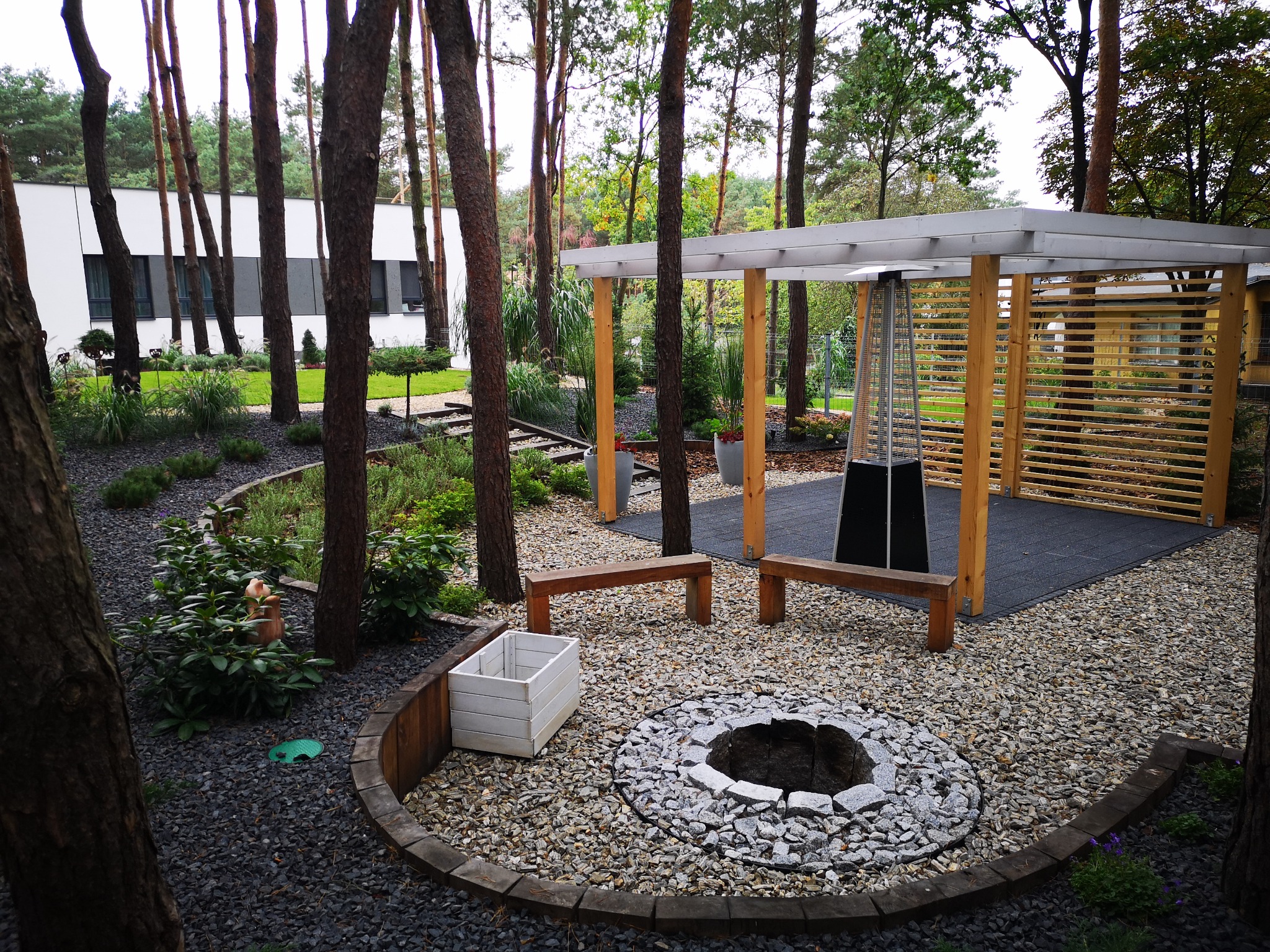Ogród z paleniskiem otoczonym kamieniami, drewnianymi ławkami i pergolą. Widoczne drzewa i nowoczesny budynek w tle. Czarna kora i roślinność.