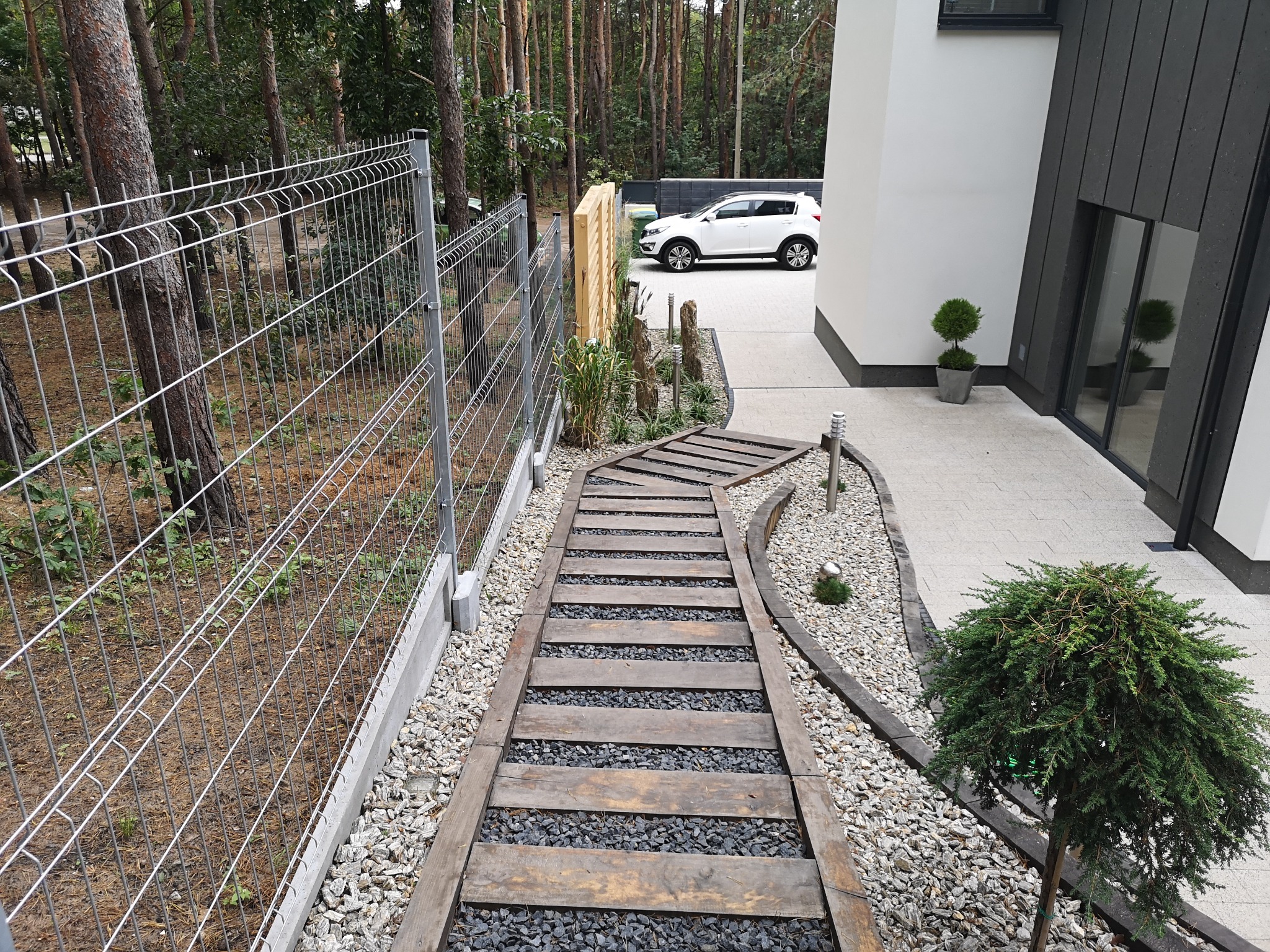 Ogród z drewnianą ścieżką ułożoną na żwirze, obok domu z białym samochodem w tle i metalowym ogrodzeniem. Dekoracyjne rośliny.