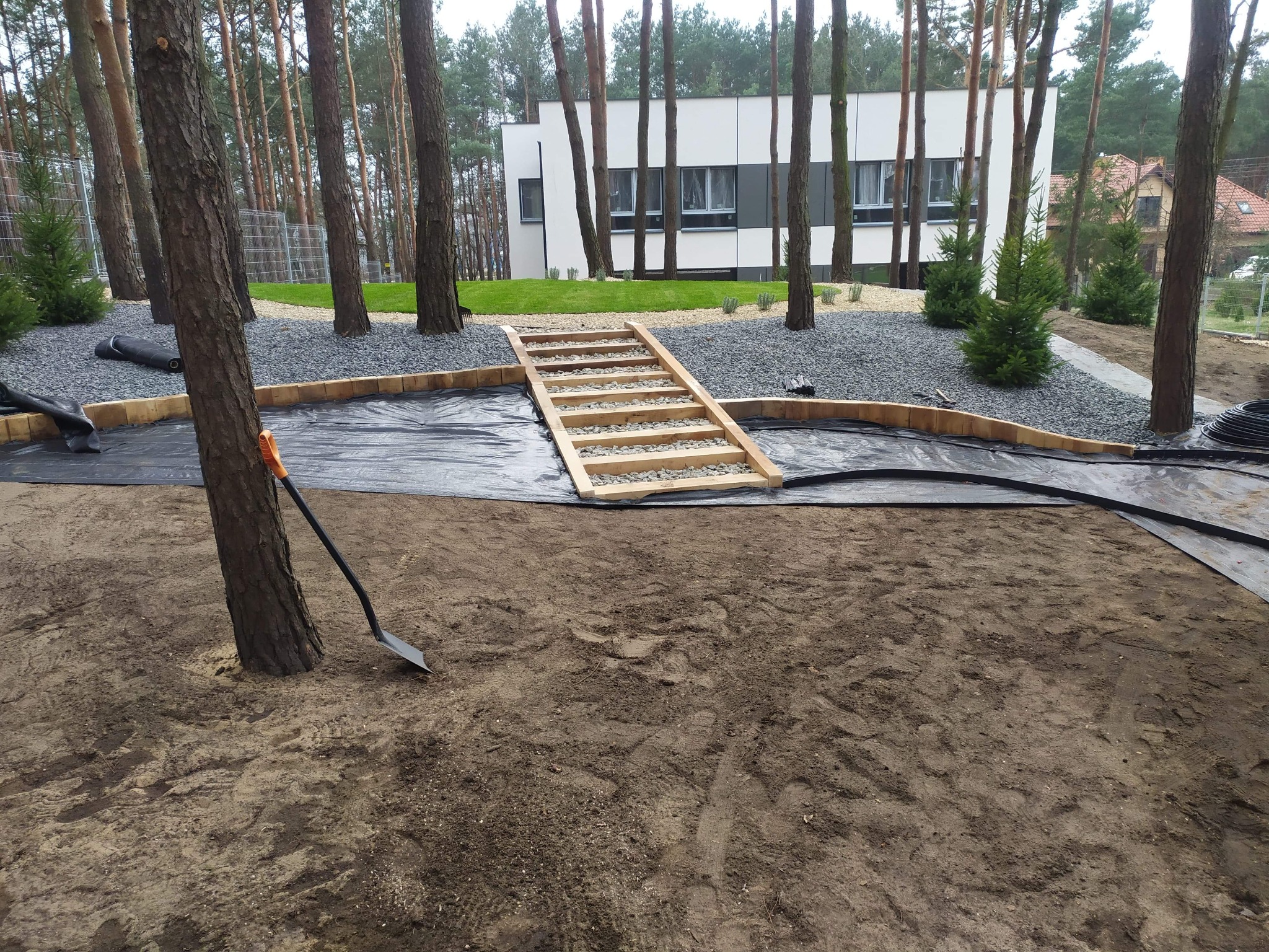 Etap zakładania ogrodu: rozłożona agrowłóknina, drewniane schodki, obrzeża z kostki, szpadel oparty o drzewo. W tle budynek i drzewa.