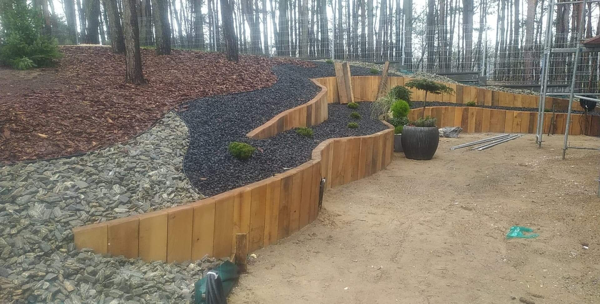 Kaskadowy ogród z drewnianymi palisadami, wypełniony czarnym grysem i roślinami, z widocznymi elementami konstrukcyjnymi w tle.