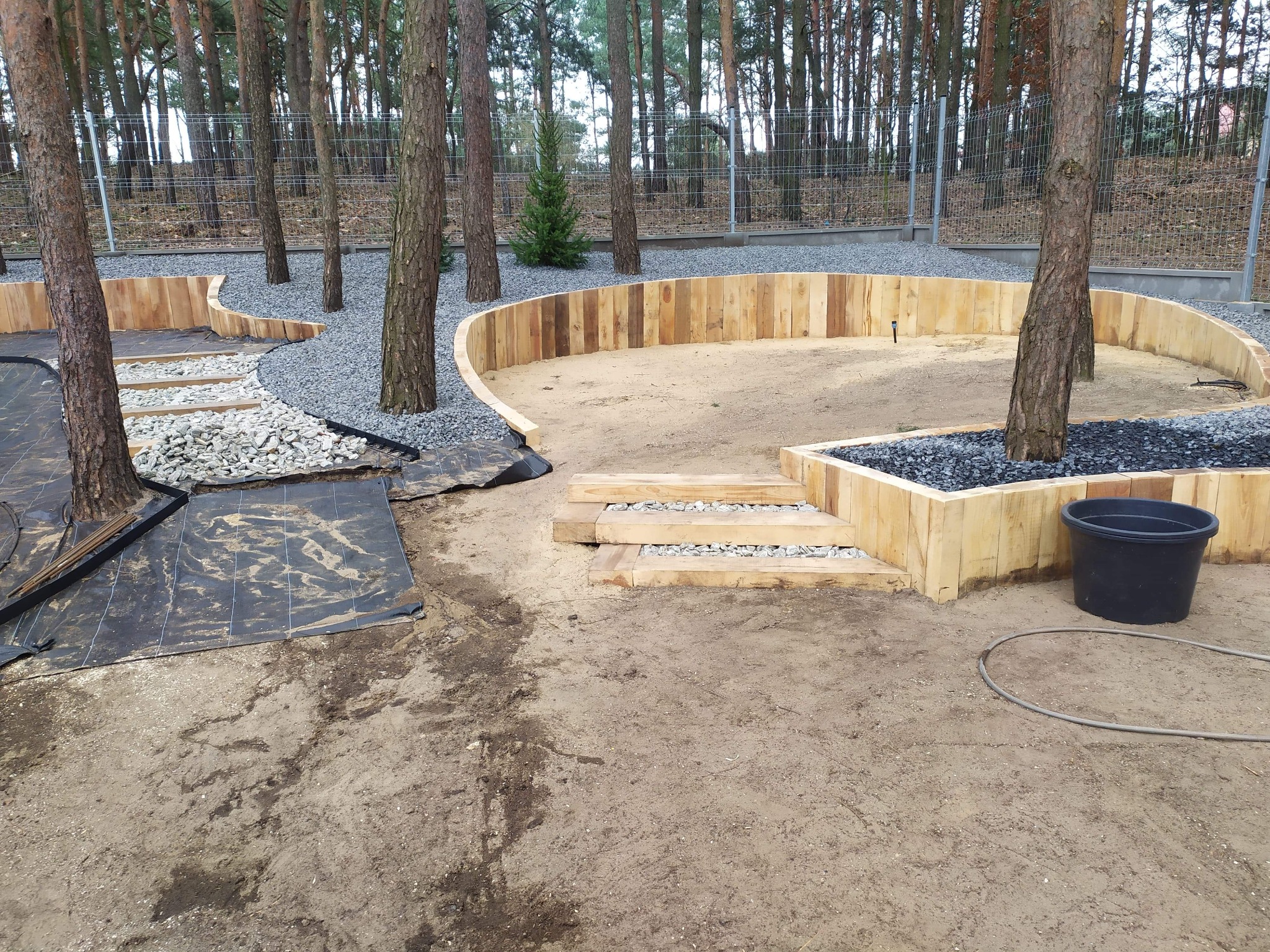 Wykonywanie drewnianej obudowy rabat w leśnym ogrodzie. Etap prac z geowłókniną, kamieniami i palisadą. Czarny kubeł i wąż ogrodowy w tle.