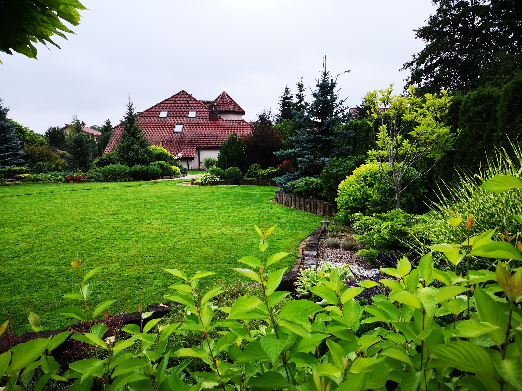 Zadbany ogród z równo przystrzyżonym trawnikiem, ozdobnymi krzewami i drzewami iglastymi na tle domu z czerwoną dachówką. Elementy drewnianej architektury ogrodowej.