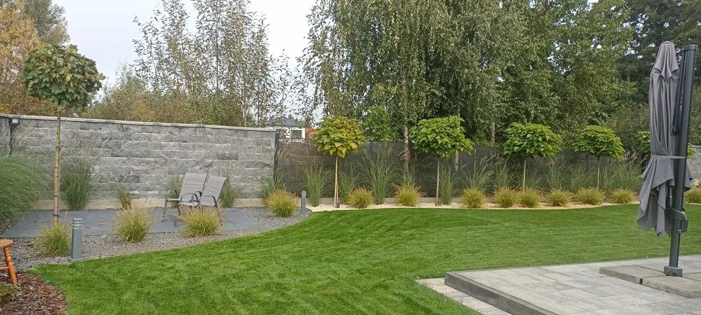 Zadbany ogród z równo przystrzyżonym trawnikiem, ozdobnymi drzewami i kamiennym murem. Uwagę przyciąga starannie zaprojektowana przestrzeń z elementami małej architektury.