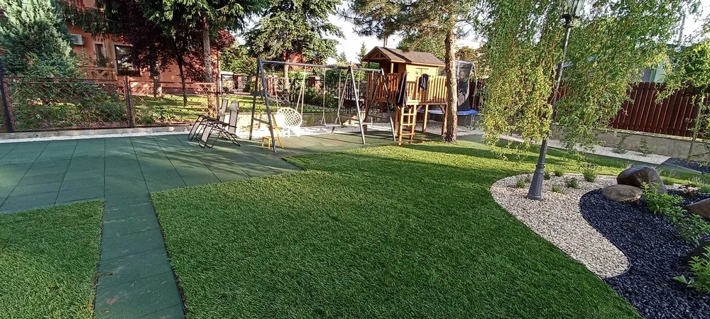 Nowoczesny plac zabaw z zieloną trawą, huśtawkami i domkiem na drzewie, otoczony ozdobnymi rabatami z kamieni i roślin. Widoczne gumowe płyty podłoża.