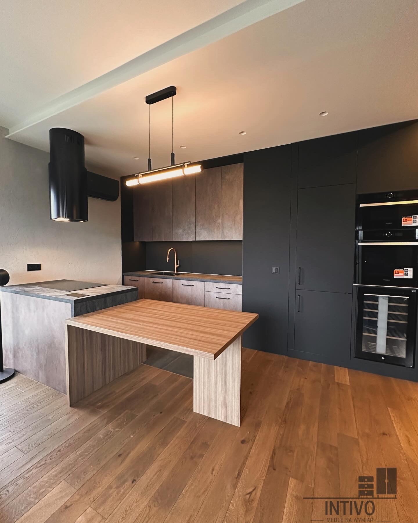 Nowoczesna kuchnia z wyspą i drewnianym blatem. Minimalistyczne szafki w kolorach szarości i czerni. Oświetlenie LED podkreśla elegancję wnętrza.