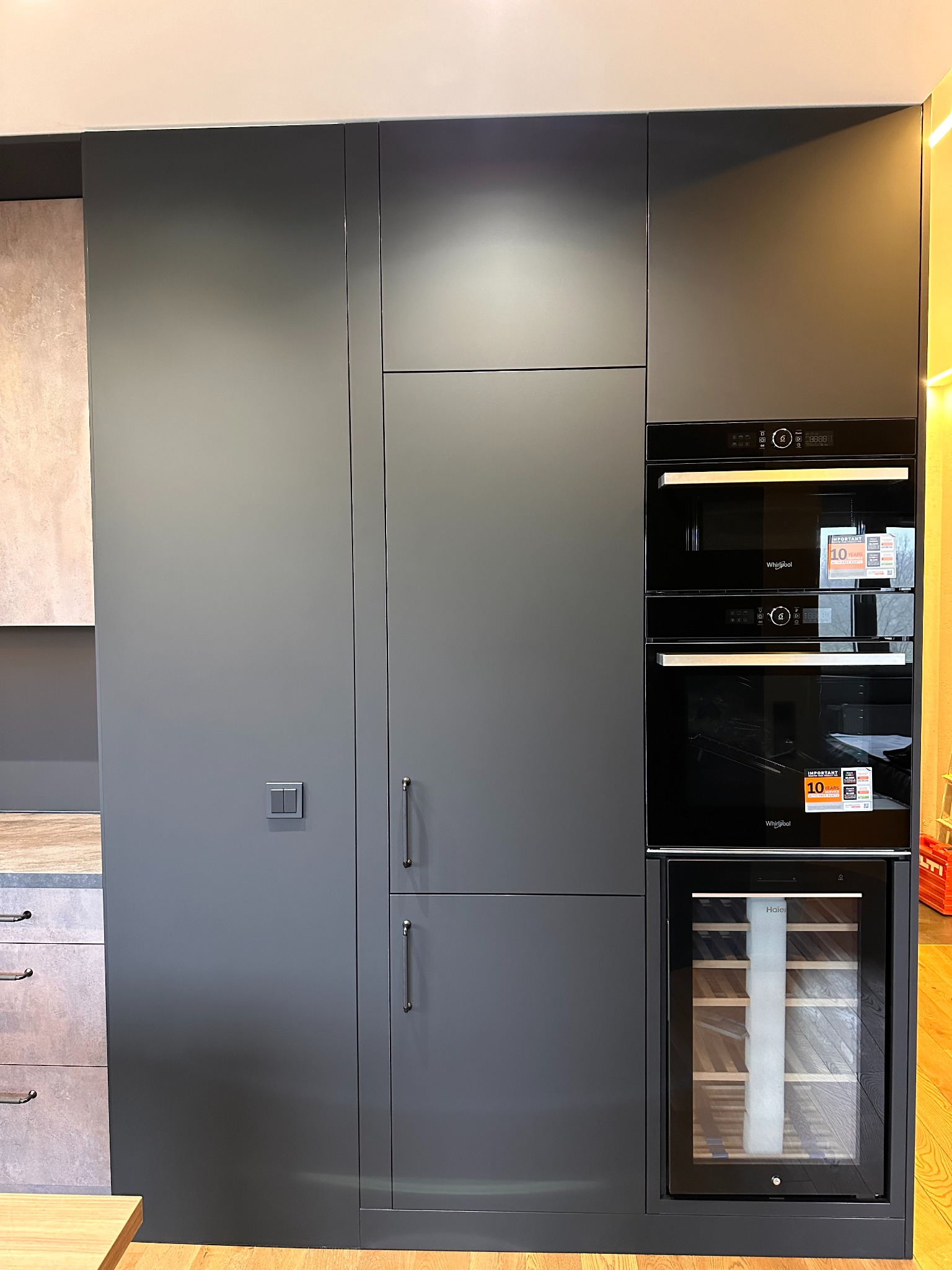 Nowoczesna, szara zabudowa kuchenna z piekarnikiem, mikrofalówką i chłodziarką do wina. Minimalistyczny design, gładkie fronty, metalowe uchwyty. Fragment blatu widoczny z lewej.