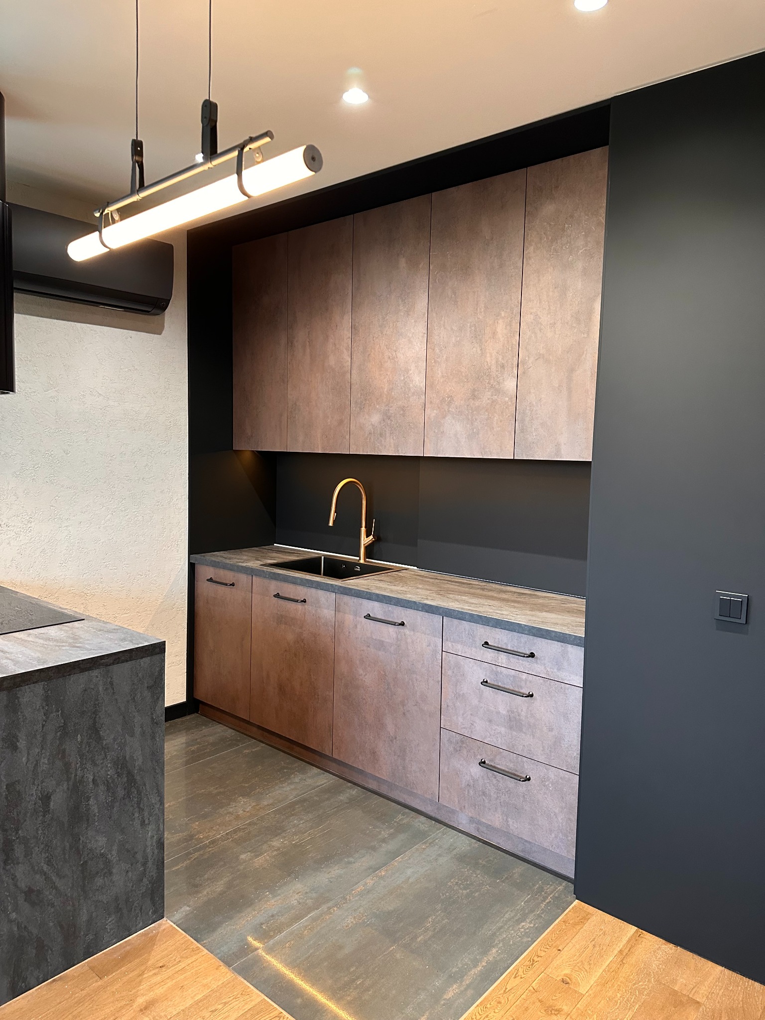 Nowoczesna kuchnia z frontami w odcieniach brązu i szarości. Złota bateria i industrialna lampa nad blatem dodają elegancji. Minimalistyczny design.