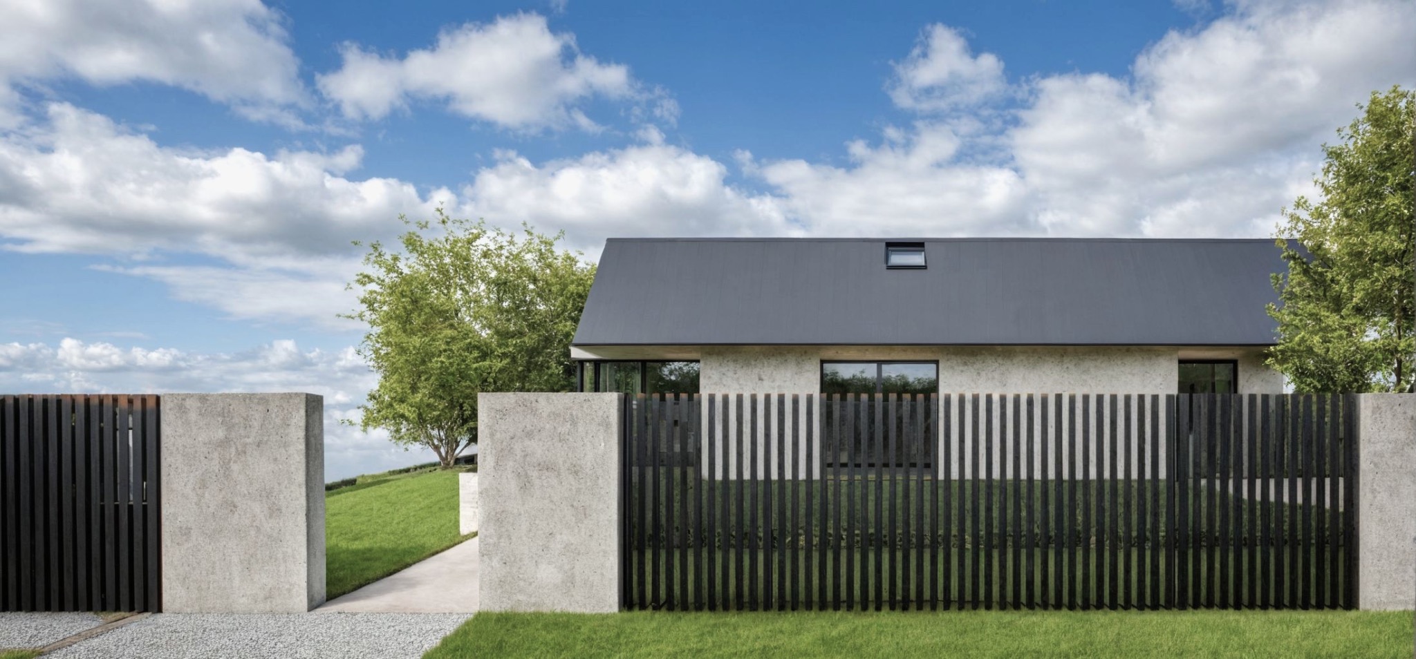 Nowoczesne ogrodzenie z ciemnych, pionowych desek i betonowych słupków na tle domu z szarą dachówką i zieleni. Minimalistyczny design, błękitne niebo z chmurami.