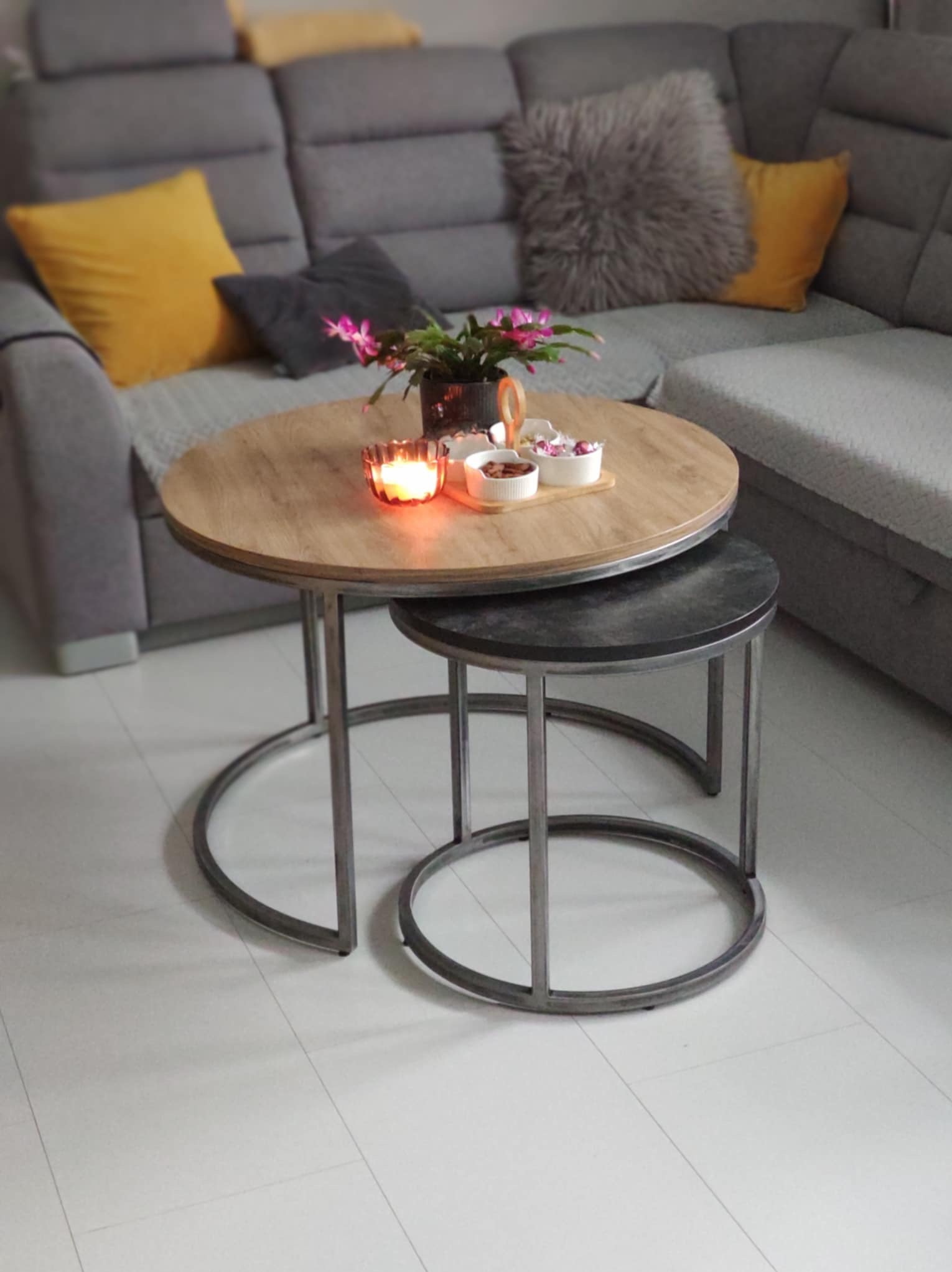 Dwa okrągłe stoliki kawowe z metalową konstrukcją, jeden większy z blatem imitującym drewno, drugi mniejszy z ciemnym blatem, ustawione na białej podłodze, w tle szara sofa z poduszkami.