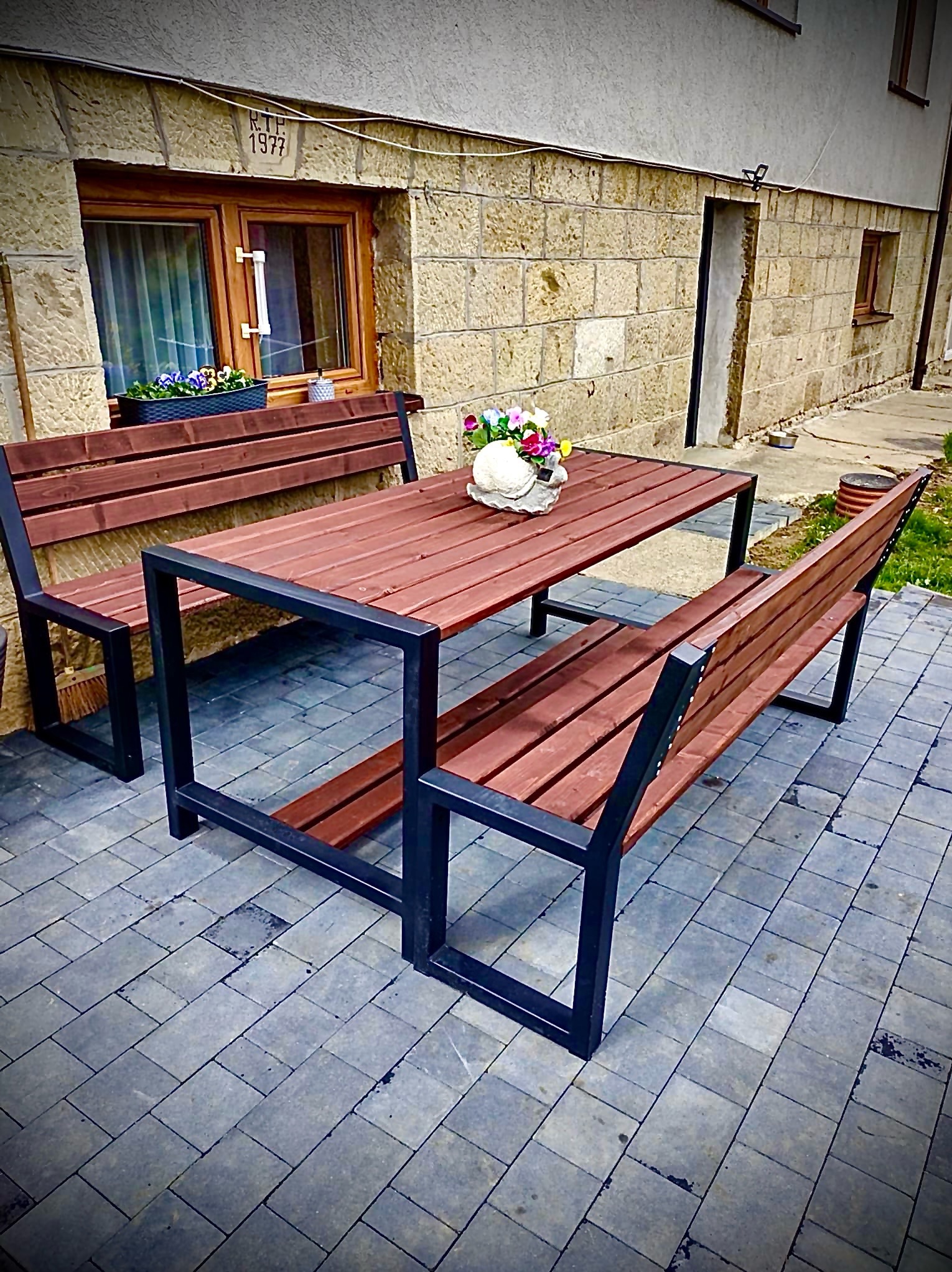 Zestaw mebli ogrodowych: stół i dwie ławki z drewnianymi siedziskami i czarną, metalową konstrukcją. Na stole dekoracja kwiatowa, w tle budynek z kamienia.