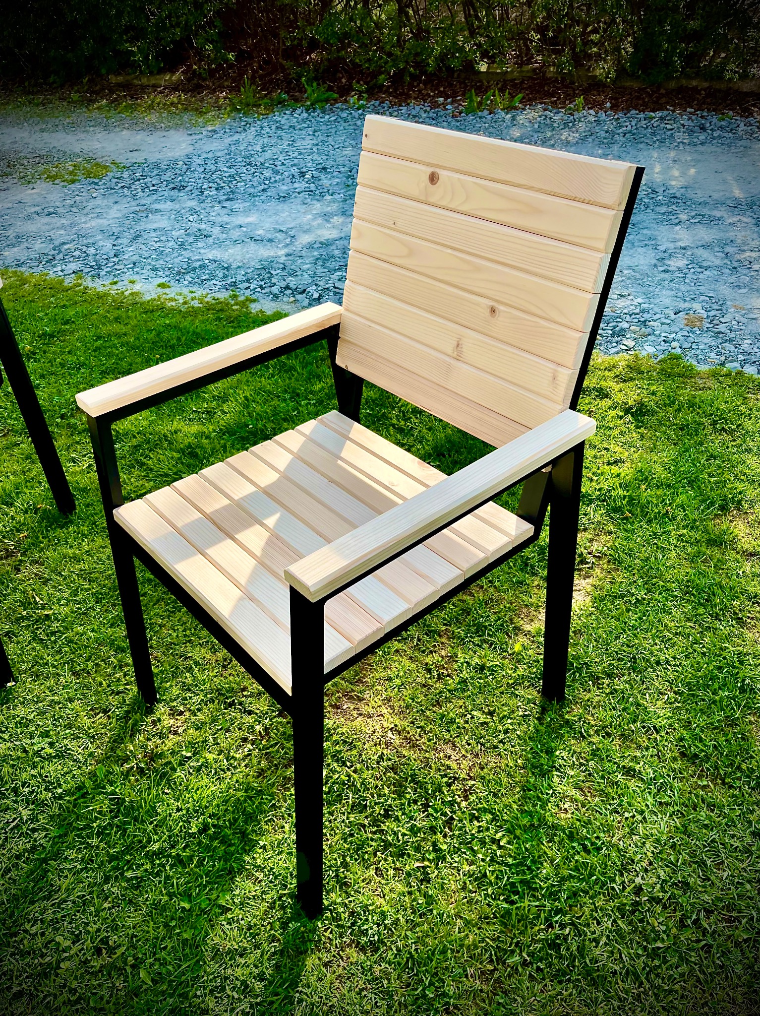 Nowoczesne krzesło ogrodowe z metalową ramą i drewnianym siedziskiem oraz oparciem, stojące na trawie. Widoczny fragment drugiego krzesła.