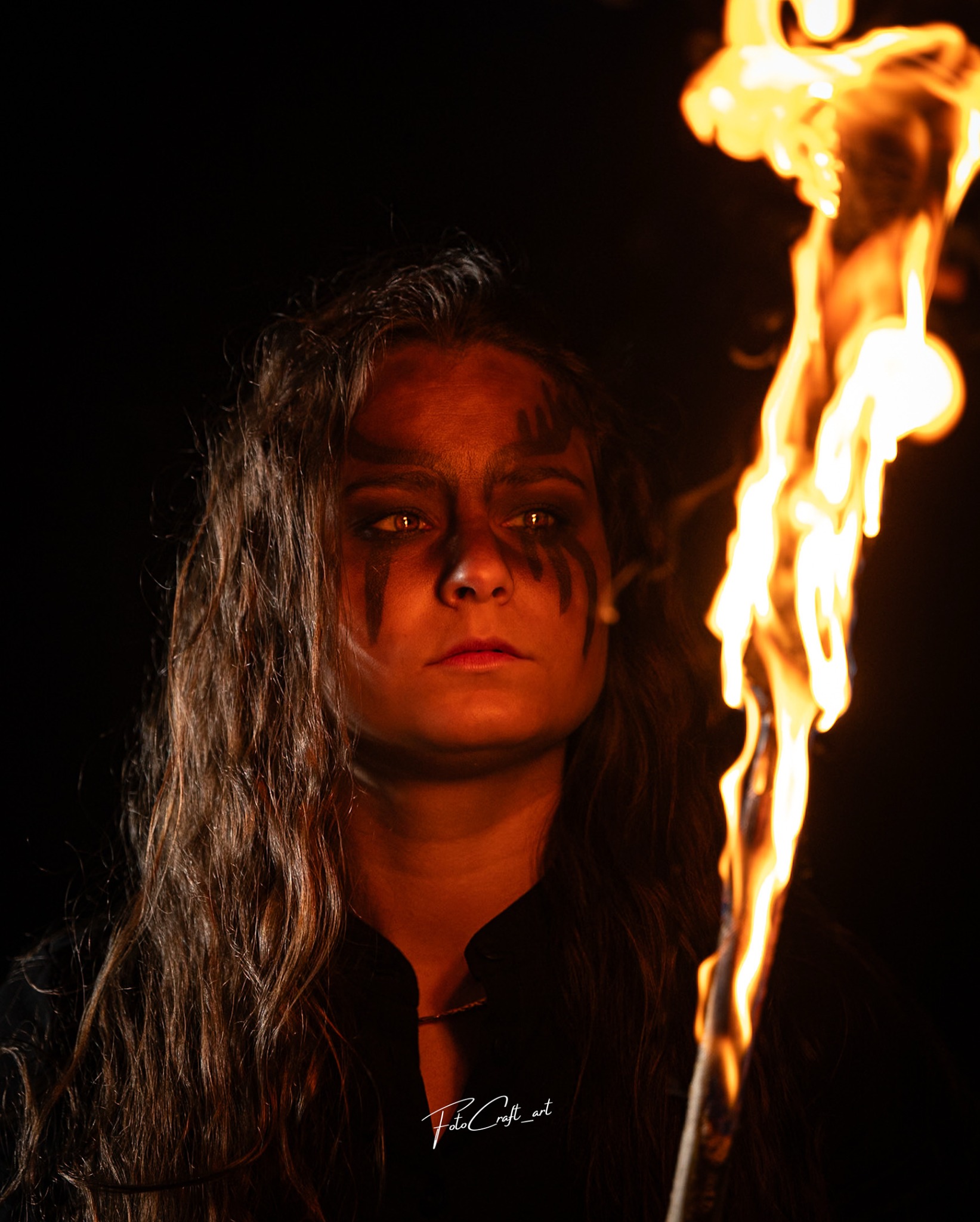 Młoda kobieta z ciemnym makijażem, trzymająca płonącą pochodnię na ciemnym tle. Wyraziste oświetlenie podkreśla ogień i rysy twarzy modelki.
