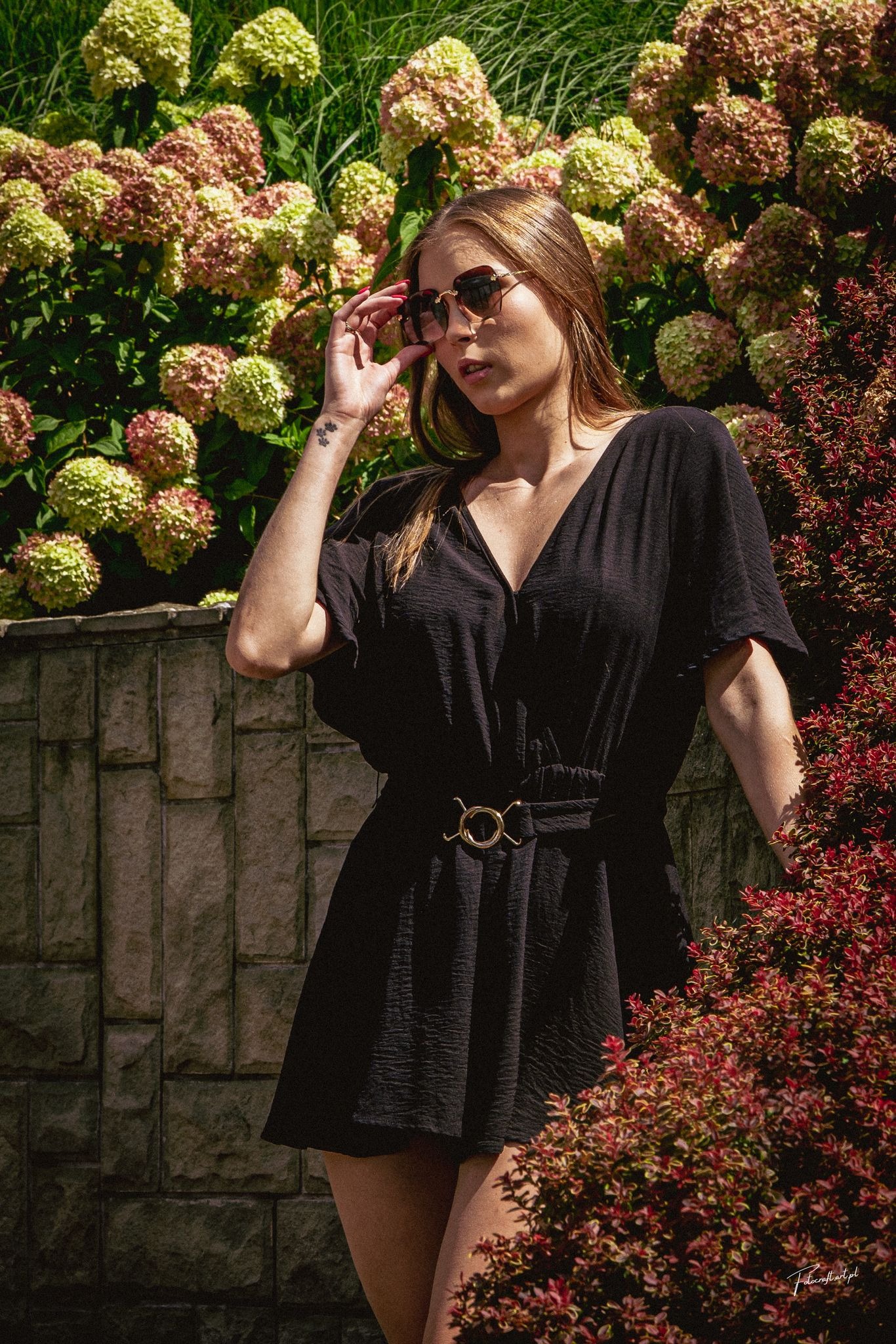 Kobieta w czarnej sukience i okularach pozuje na tle kwitnących hortensji i kamiennego murku. Widoczny tatuaż na ręce. Ujęcie w plenerze, ciepłe światło.