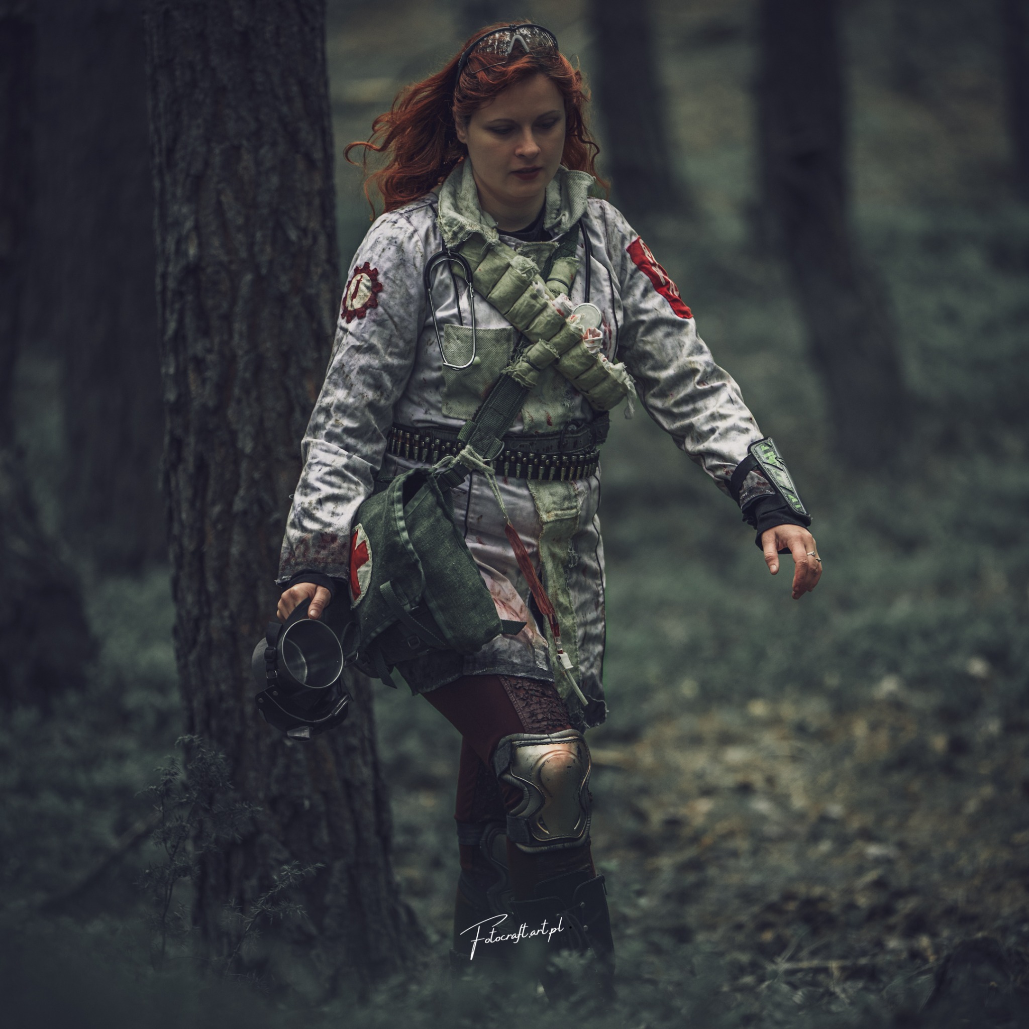 Kobieta z rudymi włosami w stylizowanej, postapokaliptycznej stylizacji medyka, idzie w lesie, trzymając maskę gazową. Ubrana w brudny, podarty kitel i wojskową amunicję.