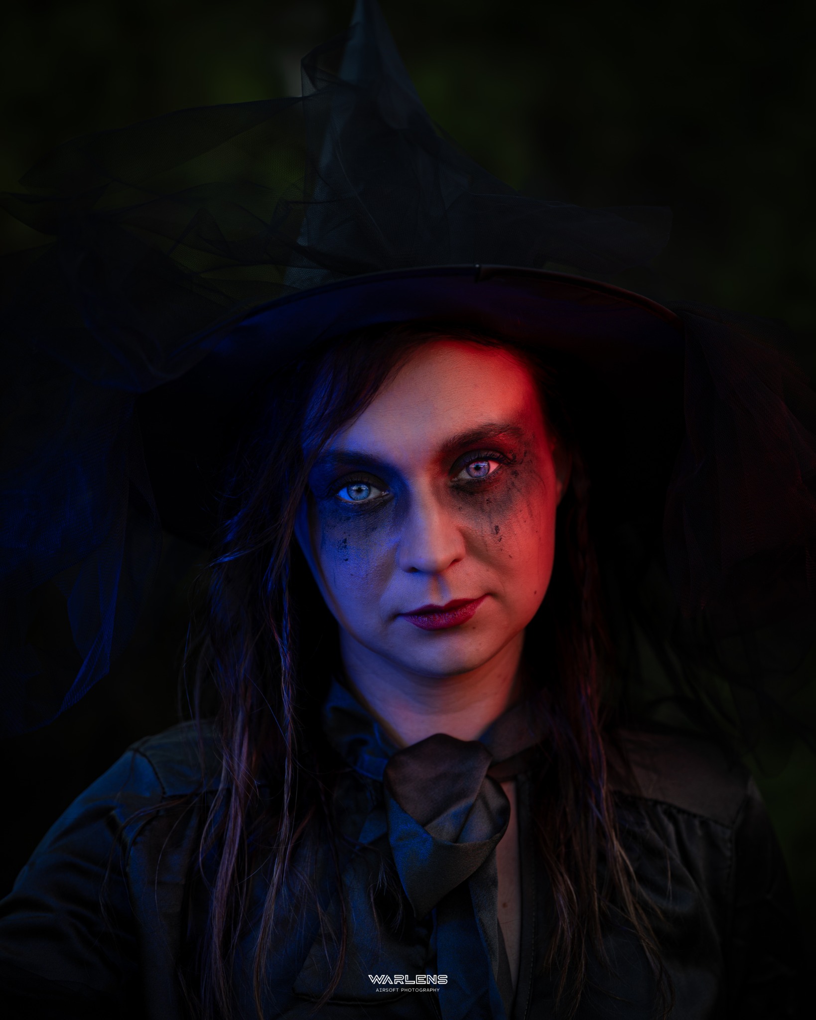 Portret kobiety w kapeluszu czarownicy, z mocnym makijażem, oświetlonej czerwonym i niebieskim światłem, na ciemnym tle. Ujęcie z bliska.