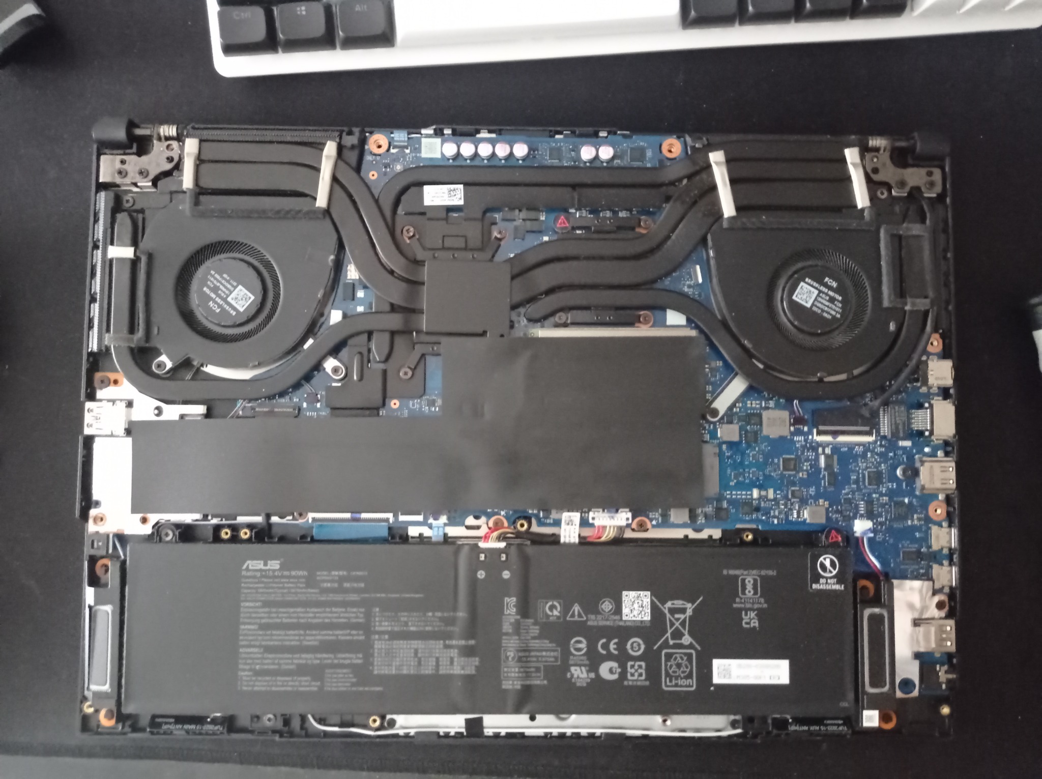 Wnętrze otwartego laptopa Asus z widoczną baterią, układem chłodzenia z dwoma wentylatorami i płytą główną, na czarnym tle klawiatura.