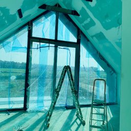 Deco-Bor Tomasz Borkowski - Poddasze w trakcie wykańczania, zabezpieczone folią okno szczytowe, dwie drabiny, widoczne ślady szpachlowania na skośnych ścianach i suficie, przewody elektryczne na podłodze.