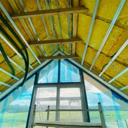 Deco-Bor Tomasz Borkowski - Skośny sufit poddasza w trakcie izolacji wełną mineralną, widoczne profile metalowe, drewniane belki, rury wentylacyjne i okno zabezpieczone folią ochronną, drabina w pomieszczeniu.