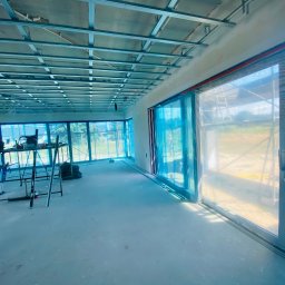 Deco-Bor Tomasz Borkowski - Montaż metalowej konstrukcji sufitu podwieszanego w przestronnym wnętrzu, pracownik na szczudłach przy rusztowaniu, okna zabezpieczone folią.