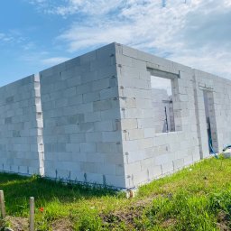 Deco-Bor Tomasz Borkowski - Surowy stan budynku w trakcie wznoszenia ścian z białych bloczków, widoczne otwory okienne i drzwiowe, teren trawiasty wokół, błękitne niebo w tle.