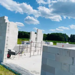 Wznoszenie murów z białych bloczków na fundamencie, widoczne rusztowanie, taczka i narzędzia, w tle zielona łąka i las pod błękitnym niebem z białymi chmurami.