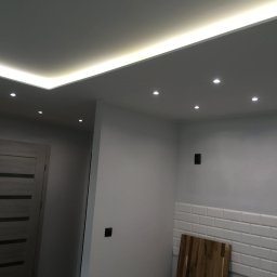 Deco-Bor Tomasz Borkowski - Nowoczesne wnętrze z podwieszonym sufitem z oświetleniem LED, punktowe lampy sufitowe, szara drewniana drzwi i fragment ściany wyłożonej białymi płytkami typu metro.