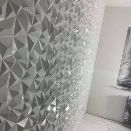 Deco-Bor Tomasz Borkowski - Ściana pokryta trójwymiarową, geometryczną strukturą z błyszczących, białych elementów, z widocznym zabezpieczeniem podłogi folią malarską i fragmentem obrazu na ścianie obok.