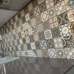 Deco-Bor Tomasz Borkowski - Ściana nad blatem kuchennym wyłożona dekoracyjnymi, kwadratowymi płytkami w stylu patchwork, utrzymanymi w odcieniach beżu, brązu, zieleni i szarości, z widocznym fragmentem zlewu i baterii.