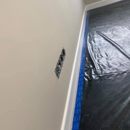 Deco-Bor Tomasz Borkowski - Ściana po nałożeniu jasnej gładzi, z widocznym gniazdkiem elektrycznym bez osprzętu, zabezpieczona folią podłoga i taśma malarska przy listwie przypodłogowej.