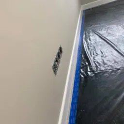 Ściana po nałożeniu jasnej gładzi, z widocznym gniazdkiem elektrycznym bez osprzętu, zabezpieczona folią podłoga i taśma malarska przy listwie przypodłogowej.