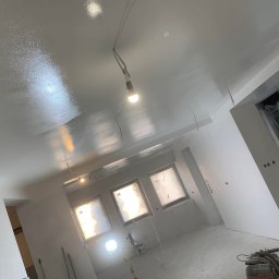 Deco-Bor Tomasz Borkowski - Świeżo położona, błyszcząca gładź na suficie w trakcie schnięcia, widoczne tymczasowe oświetlenie, zakryte okna i sprzęt do szlifowania na podłodze.