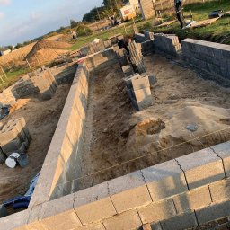 Deco-Bor Tomasz Borkowski - Budowa fundamentów domu jednorodzinnego z bloczków betonowych, widoczny wykop, ułożone pierwsze warstwy bloczków, poziomowanie sznurkiem, zapas bloczków na paletach i wiaderka z zaprawą wewnątrz...