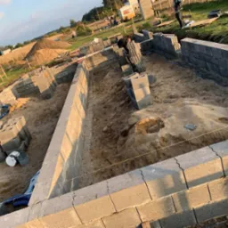 Budowa fundamentów domu jednorodzinnego z bloczków betonowych, widoczny wykop, ułożone pierwsze warstwy bloczków, poziomowanie sznurkiem, zapas bloczków na paletach i wiaderka z zaprawą wewnątrz...