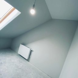 Deco-Bor Tomasz Borkowski - Poddasze po świeżym malowaniu na szaro, widoczny grzejnik pod oknem dachowym i surowa podłoga przed wykończeniem, pojedyncza żarówka zwisa z sufitu.