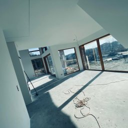 Deco-Bor Tomasz Borkowski - Wnętrze nowo wykończonego, jasnego pomieszczenia z dużymi oknami z drewnianymi ramami, widoczne ślady po pracach wykończeniowych na podłodze i zwisający przewód elektryczny, ściany pomalowane...