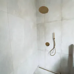 Wykończona kabina prysznicowa z dużymi, jasnymi płytkami, złotą armaturą i odpływem liniowym w podłodze.