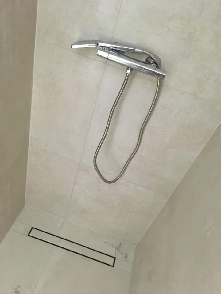 Nowoczesna kabina prysznicowa z odpływem liniowym i chromowaną baterią natryskową na tle jasnych, kwadratowych płytek ceramicznych.