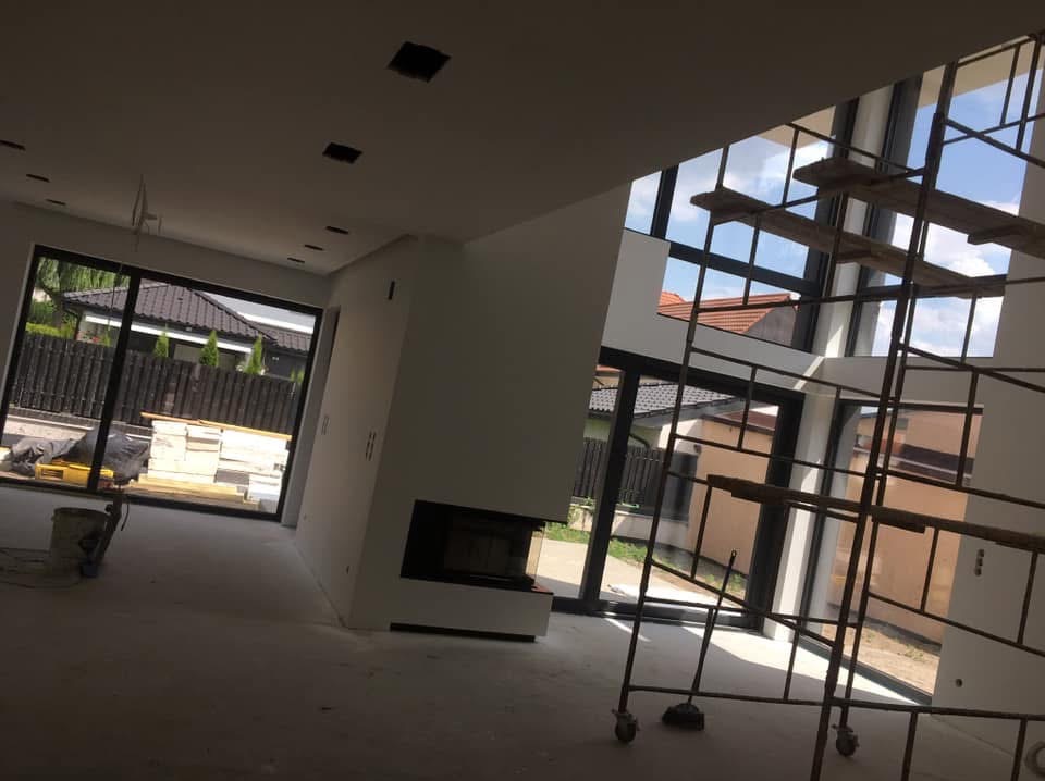 Wnętrze w trakcie wykańczania: ściany przygotowane pod malowanie, widoczne rusztowanie przy oknie, surowa podłoga, nowoczesny kominek w zabudowie.