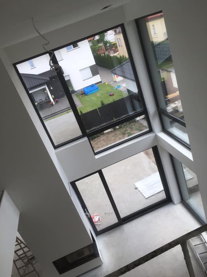 Wnętrze domu w trakcie wykańczania z dużymi oknami z widokiem na posesję z ogrodem, fragment ściany z kominkiem, surowe ściany i podłoga.