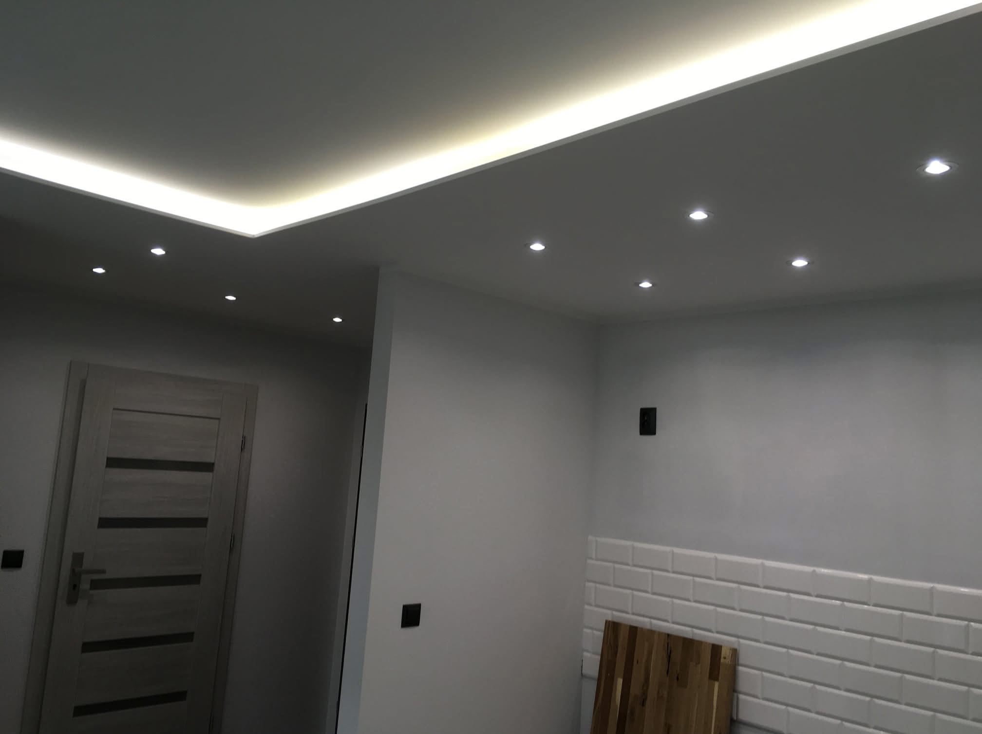 Nowoczesne wnętrze z podwieszonym sufitem z oświetleniem LED, punktowe lampy sufitowe, szara drewniana drzwi i fragment ściany wyłożonej białymi płytkami typu metro.