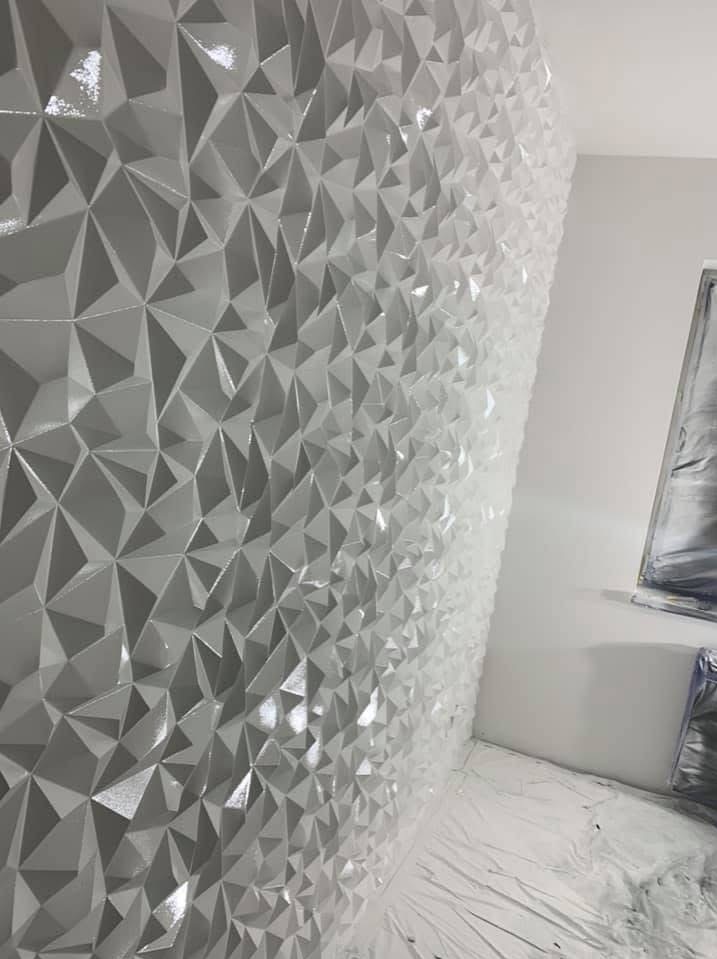 Ściana pokryta trójwymiarową, geometryczną strukturą z błyszczących, białych elementów, z widocznym zabezpieczeniem podłogi folią malarską i fragmentem obrazu na ścianie obok.