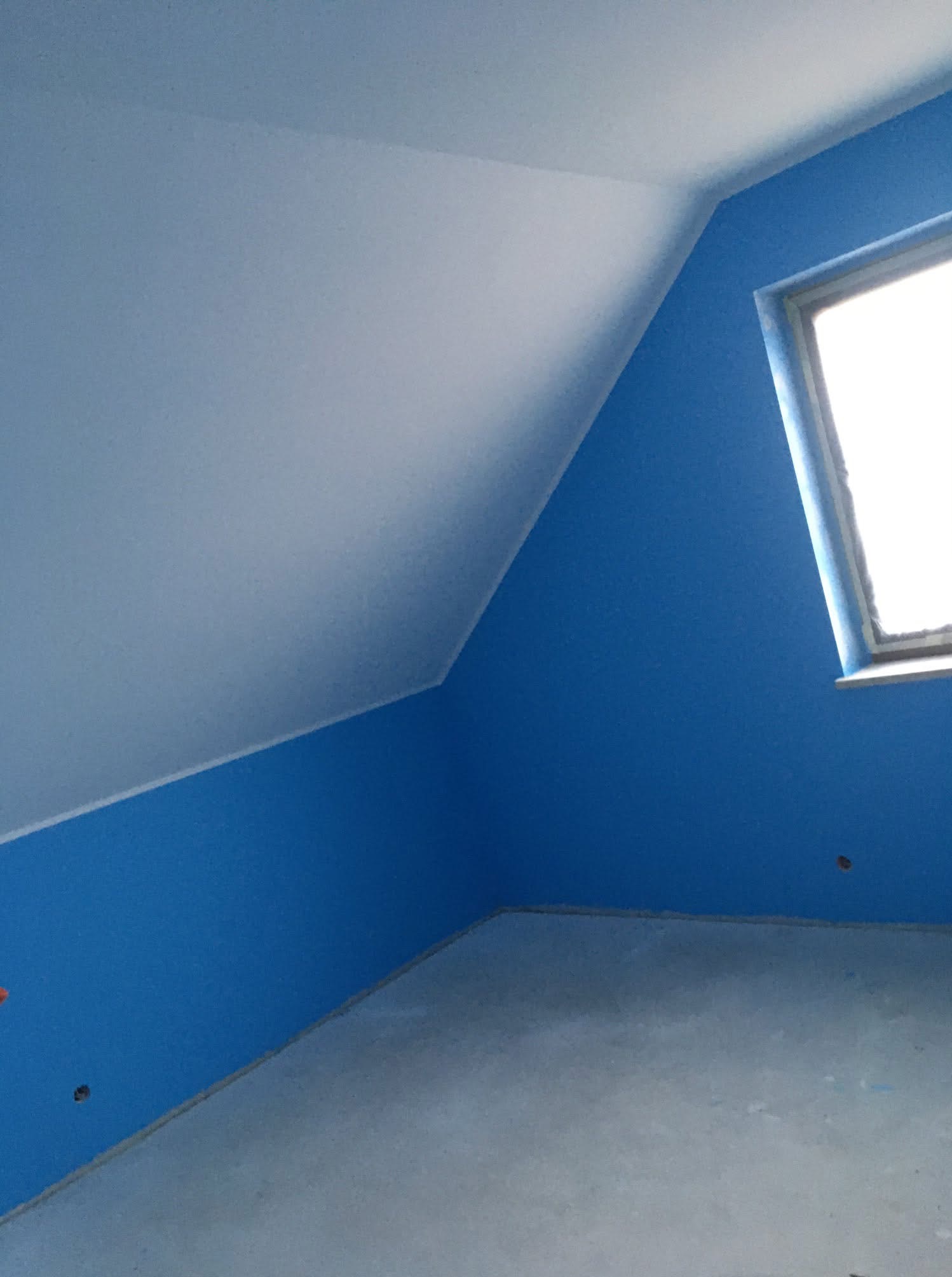 Pokój na poddaszu z pomalowanymi na niebiesko ścianami, biały sufit ze skosem, okno z taśmą malarską na framudze, widoczne niedoskonałości wykończenia przy podłodze.