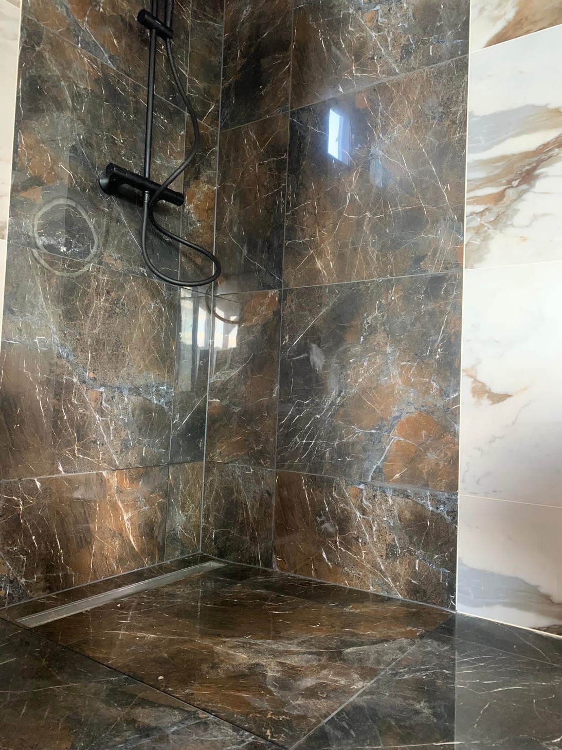 Nowoczesny prysznic walk-in z czarną armaturą i ciemnobrązowymi płytkami imitującymi marmur z białymi żyłami, kontrastującymi z jasnymi płytkami na sąsiedniej ścianie.